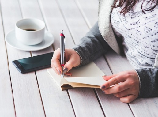 Téli női kéz ír egy kerti asztalon, mellette a telefonja, és egy csésze kávé, Kép: pixabay